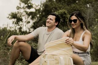 A smiling couple enjoys a relaxed moment outdoors, surrounded by greenery, with a picnic bag beside them. They appear cheerful and carefree.