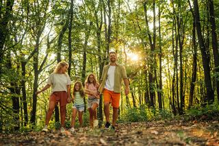 A family walks hand-in-hand through a sunlit forest, surrounded by tall trees and autumn foliage, enjoying a joyful outdoor moment.