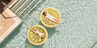 Two women relax on large lemon-shaped floaties in a sparkling pool, enjoying a sunny day. Steps lead down to the water's edge.