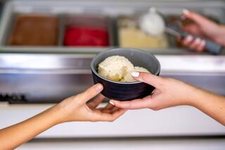 A hand is passing a bowl of ice cream, while another hand holds a scoop near a display of various frozen treats.