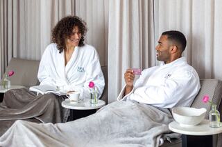 A woman and a man in cozy robes relax together, enjoying drinks and conversation in a serene spa-like environment.