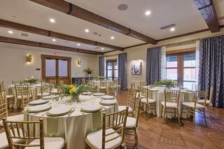 Elegant event space with round tables dressed in cream linens and gold chairs, adorned with fresh flowers, and large windows for natural light.