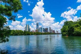 A vibrant city skyline reflects in a serene lake, surrounded by lush greenery and fluffy clouds in a bright blue sky.