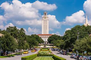 A grand clock tower stands tall amidst lush greenery, with a pathway lined by trees and parked cars, under a bright blue sky with fluffy clouds.