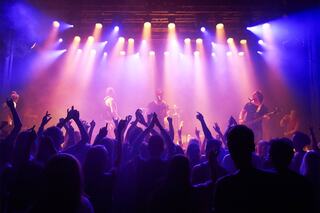 A lively crowd enjoys a concert, hands raised in excitement, illuminated by vibrant stage lights. Musicians perform passionately.