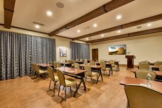 A spacious meeting room features wooden beams, tables with chairs, and a podium, complemented by dark curtains and a decorative wall.