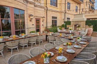 A beautifully set outdoor dining area features a long wooden table adorned with flowers and dishes, surrounded by stylish chairs and greenery.