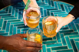 Three hands hold glasses of golden drink with ice, garnished with citrus, against a vibrant teal-patterned table. Cheers!