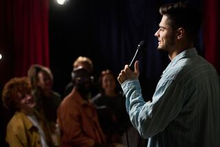 A performer stands on stage holding a microphone, engaging an amused audience in a lively comedy club atmosphere.