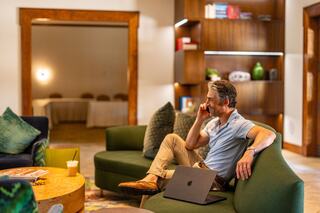 A casually dressed man sits on a green sofa, talking on his phone with a laptop nearby, surrounded by a cozy, modern interior.