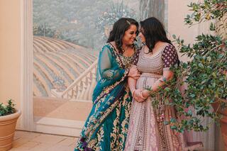 Two women in vibrant traditional attire share a joyful moment, standing together amidst lush greenery and a scenic backdrop.