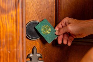 A hand holds a key card near a door lock, preparing to gain access, with a wooden door showcasing intricate details.