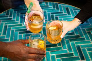 Three hands hold glasses of cocktails with ice and citrus, set on a vibrant teal tiled table, celebrating togetherness.