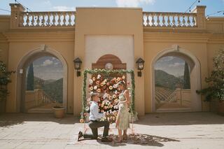 A man kneels in front of a woman against a floral backdrop, capturing a moment of proposal in a beautiful outdoor setting.