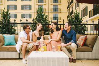 Four friends enjoy drinks while relaxing on a stylish couch, chatting and laughing in a sunlit outdoor setting.