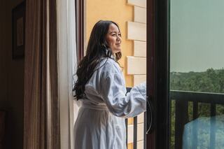 A woman in a white robe stands by an open balcony door, gazing thoughtfully at the scenic view outside.
