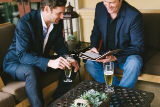 Two men sit together, enjoying drinks and discussing a menu, surrounded by a cozy outdoor setting with plants and lanterns.