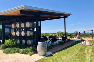 A modern winery features a patio with barrels, seating, and a scenic vineyard in the background, where visitors enjoy the outdoors.