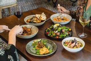 A table features various delicious dishes, including salads, pasta, and seafood, complemented by glasses of red and white wine.