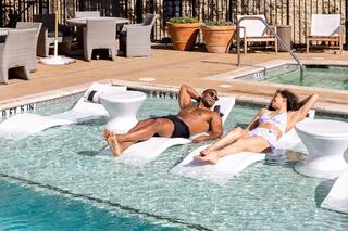 Two individuals relax on pool loungers in shallow water, enjoying a sunny day at a stylish poolside setting.