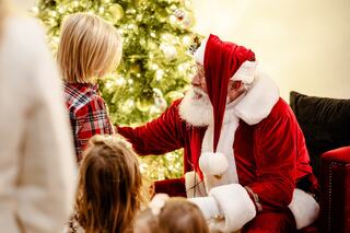 A jolly Santa in a red suit interacts warmly with a young child in front of a beautifully decorated Christmas tree.