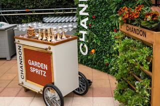 A stylish cart labeled "GARDEN SPRITZ" with elegant glasses on top, surrounded by greenery and a chic display of flowers.