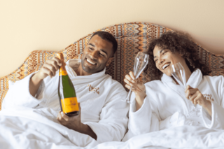 A joyful couple in bathrobes celebrates in bed, holding a champagne bottle and glasses, sharing laughter and a cozy moment.