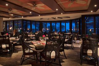 A dimly lit restaurant features elegant seating and floral centerpieces, with large windows showcasing a scenic view of the evening sky.