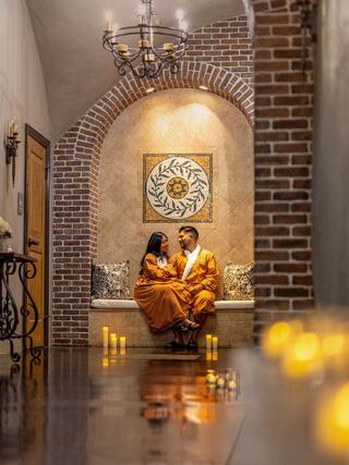 A couple in matching orange robes shares an intimate moment in a cozy, warmly lit hallway adorned with candles and a decorative wall.