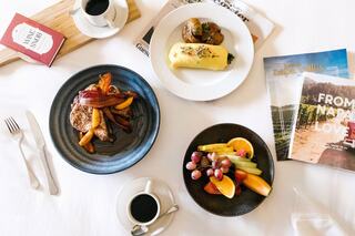 A stylish breakfast spread features a savory dish with vegetables, a sweet pastry, fresh fruit, and coffee, accompanied by magazines.
