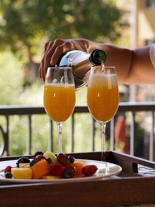 A hand pours sparkling juice into two flutes next to a plate of colorful fruit on a sunny balcony.