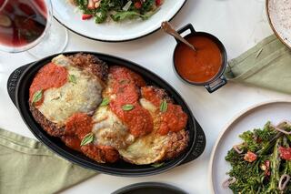 Crispy chicken covered in marinara sauce and melted cheese, garnished with fresh herbs, alongside a side of salad and a glass of wine.