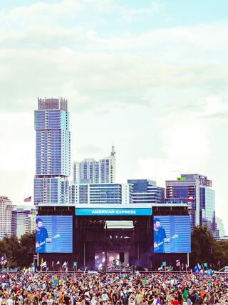 A lively outdoor concert scene features a large crowd in front of a stage, with skyscrapers and a clear sky in the background.