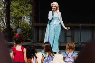 A performer dressed as a princess sings on stage, entertaining a group of young children dressed in colorful costumes, enjoying the show.