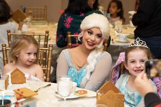 A smiling woman with long, blonde hair, dressed as a princess, interacts with two young girls in costumes at a festive table.
