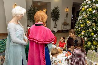 Two characters dressed as princesses interact with children at a festive table, decorated for a holiday celebration with a Christmas tree nearby.