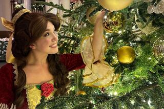 A young woman in a festive dress decorates a Christmas tree with gold ornaments, smiling joyfully amidst twinkling lights.
