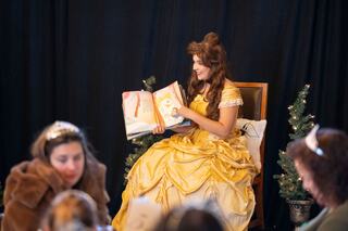 A storyteller in a golden gown reads a book, while an audience of children listens intently amid festive decorations.