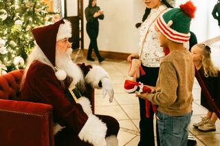 A child hands a gift to Santa Claus, while others look on, surrounded by festive decor and a Christmas tree.
