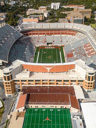 Aerial view of a large stadium with green football field and seating, featuring the Texas Longhorns branding and nearby buildings.