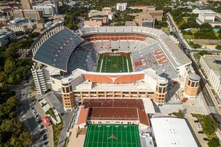 Aerial view of a large stadium surrounded by urban buildings, featuring a green football field and the iconic Texas Longhorns logo.