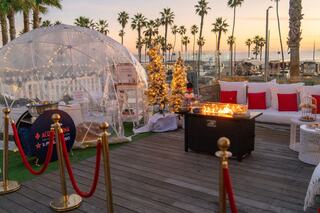A cozy outdoor space features a transparent dome, festive decor, a fire pit, and palm trees against a sunset backdrop.