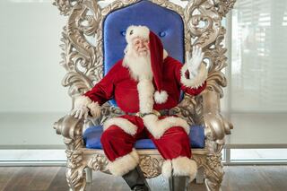 A jovial Santa Claus in a red suit sits regally on an ornate silver throne, waving warmly with a bright smile.