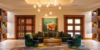 A cozy lounge features a green sofa, vibrant patterned chairs, wooden accents, and warm lighting, creating an inviting atmosphere.