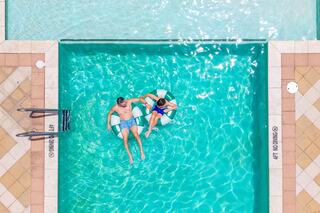 A couple relaxes on floaties in a shimmering pool, surrounded by tiled edges and a ladder, enjoying a sunny day.