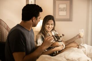 A couple enjoys a cozy moment in bed with their small dog, sharing smiles and a drink while relaxing together.