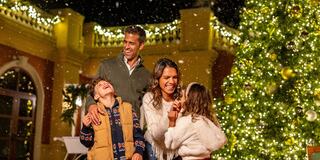 A joyful family stands under snowfall, surrounded by festive lights and a beautifully decorated Christmas tree, embracing the holiday spirit.