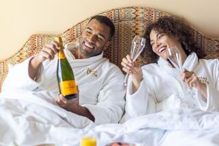 A joyful couple in cozy robes celebrates with champagne in bed, smiling and toasting with glasses, enjoying a special moment together.