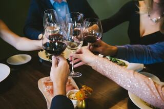 Glasses filled with wine are raised in a toast among friends, surrounded by plates of food, creating a festive dining atmosphere.