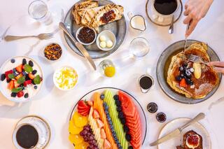 A vibrant brunch spread featuring pancakes, fresh fruit, yogurt, and coffee, with hands serving and enjoying the delicious meal.